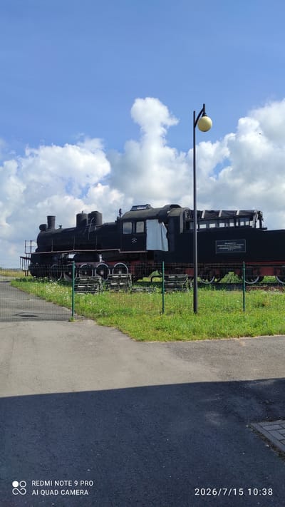 This image shows a black steam locomotive on display, parked on a track surrounded by green grass and a low fence. The locomotive is a large, old-fashioned train with multiple wheels and a tall smokestack. A modern street lamp with a round, yellow light is positioned to the right of the train. The background consists of a bright blue sky with scattered white clouds. A plaque is visible on the side of the locomotive. The scene appears to be an outdoor exhibit or museum. The image is dated 2026/7/15 10:38 and includes a watermark indicating it was taken with a REDMI NOTE 9 PRO AI QUAD CAMERA. The location specified is Лашанскі сельскі Савет, Belarus.