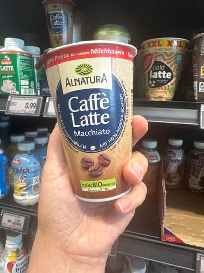 The image shows a hand holding a cup of Alnatura Caffé Latte Macchiato. The cup is beige with blue and brown text and graphics, including images of coffee beans. The background consists of shelves in a grocery store, stocked with various food and beverage items, including other coffee drinks and milk-based products. Some of the visible labels include "Alnatura", "XXL", "M Hijo", "Milch", and "Bio". The price tag "0.99" is visible on a shelf in the lower left corner. The scene appears to be in a supermarket, possibly in Tuntenhausen, Germany, given the presence of German text on the product packaging. The lighting is artificial, consistent with indoor retail lighting. The focus is on the Alnatura cup, with the background slightly blurred.