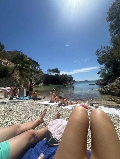 The image depicts a sunny day at a beach in Peguera, Spain. The beach is crowded with people sunbathing and swimming in the water. The shoreline is composed of small stones. The foreground focuses on the legs of an individual lounging on a blue towel, a light pink bag sitting nearby. Several people can be seen in the background, swimming in the water and sunbathing on towels. The sky is clear with some wispy clouds. A rocky cliff and trees are visible along one side of the beach, with a building nestled among the trees. A boat is seen floating on the water.