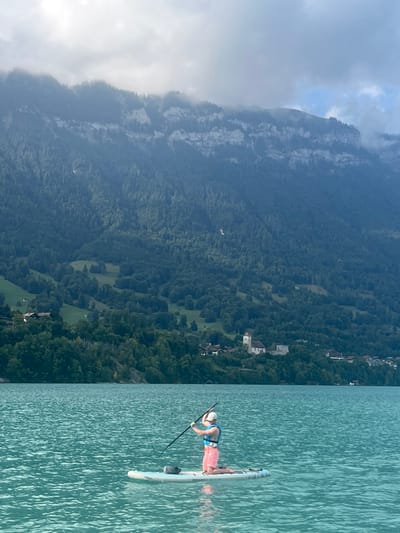 An individual is depicted kneeling on a light-colored stand-up paddleboard on the turquoise waters of Lake Brienz, in Bönigen, Switzerland. The person wears a white baseball cap, a light blue life vest over a light-colored top, and pink-patterned shorts, holding a black paddle with both hands in a ready or mid-stroke position. A dark object, possibly a dry bag, rests on the front of the paddleboard. The background features the shoreline of Bönigen with a prominent light-colored church, distinguished by its steeple, among other buildings and lush green trees. Behind the village, steep, heavily forested mountains rise dramatically, exhibiting lighter rock faces and partially obscured peaks by low-hanging clouds under an overcast sky.