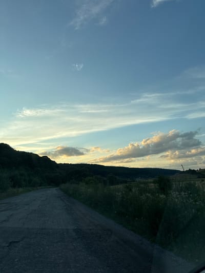 The image shows a view of a rural road leading through a landscape in the city of Rohachyn, Ukraine. The road is unpaved and appears to be made of gravel and dirt, with some visible cracks and imperfections. The road curves to the left and disappears into the distance. On the left side of the road, there is a dense, dark green forest that covers a hillside. To the right of the road, there are fields and vegetation, with some taller grasses and plants. In the background, there are rolling hills and more distant fields, some of which are illuminated by the soft light of the setting sun. The sky is a clear blue with scattered wispy clouds, some of which are tinged with yellow and orange from the sunset. The overall impression is one of a serene, natural landscape at dusk. There are no people or animals visible in the image. The scene appears to be tranquil and undisturbed.