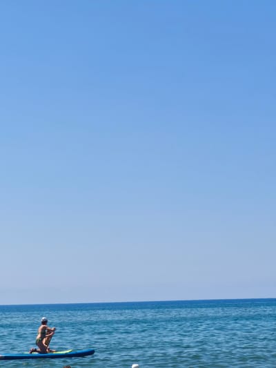 A wide shot captures a clear blue sky and the deep blue sea. In the foreground, a woman wearing a white cap and green bikini is kneeling on a blue paddleboard. She is holding a paddle and appears to be in motion on the water. To her right, a smaller figure, likely a child, is also on the paddleboard, facing away from the camera. The water is calm, with small ripples. The scene is set in the sea off the coast of Castiglione della Pescaia, Italy, a location known for its beaches and marine activities.