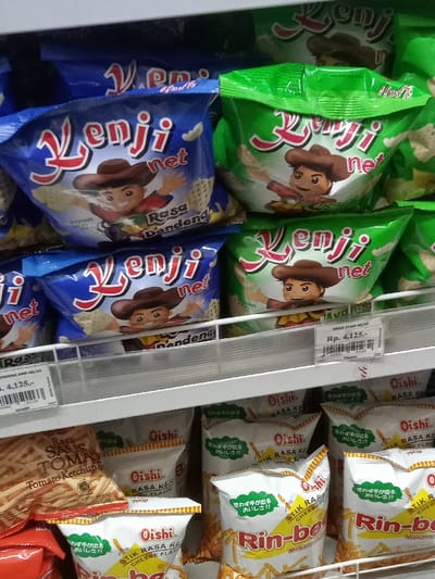 The image displays a close-up view of a retail snack food aisle within a store in Nganjuk, Indonesia. Two primary shelves are visible, both stocked with packaged snack products.
On the upper shelf, multiple bags of "Kenji net" brand snacks are arranged. On the left side, several blue bags are visible, labeled "Kenji net Rasa Dendeng" (Beef Jerky Flavor). Each bag features a cartoon depiction of a boy wearing a brown cowboy hat, with his arms outstretched, against a background of waffle-patterned snacks. A price tag on the left, partially obscured, indicates "Rasa Dendeng" and a price of Rp. 4,125,-. On the right side of the upper shelf, green bags of "Kenji net" snacks are present, also featuring the same cowboy-hat-wearing cartoon character. A clearer price tag on the right identifies these as "KEMJI AYAM 40/24" (Chicken Flavor) and also priced at Rp. 4,125,-.
The lower shelf contains multiple white bags of "Oishi Rin-bee Stik Rasa Keju" (Cheese Flavor Sticks). These bags prominently display "Oishi" and "Rin-bee" in red text, alongside Japanese characters "思わず手がでるおいしさ!!" (Unintentionally delicious!!). The bags are filled with elongated, stick-shaped snacks. To the far left of the lower shelf, a partially visible red and orange bag from the "Oishi" brand is present, labeled "Rasa Saus Tomat" (Tomato Ketchup Flavor), containing different, possibly cracker-like, snacks.
The snacks are displayed on white-colored, multi-tiered shelving units. No individuals are present in the frame, and no specific actions or interactions are depicted. The lighting is bright and even, consistent with an indoor retail environment.