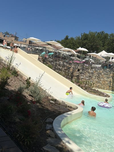 In Cavriglia, Italy, a bright, sunny day is captured at a water park. A wide, cream-colored water slide dominates the foreground, with a young boy in neon yellow swim trunks mid-slide at the bottom, splashing into a clear turquoise pool. To the right of the slide's end, a girl with blonde hair is partially submerged in the pool, and further right, a boy wearing a striped inner tube floats on his back. Another boy is visible wading in the pool. The pool area features a curved white edge, contrasting with the rocky, landscaped embankment on the left, which is dotted with green shrubs and red flowers. In the background, a stone wall supports an elevated area with lounge chairs and numerous white umbrellas providing shade. Several people are visible relaxing on the lounge chairs. The sky above is a vibrant, clear blue.