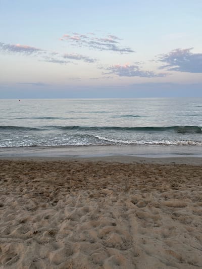 This is an eye-level, full shot of a beach scene in Grao, Spain, likely taken during dusk or dawn due to the soft lighting and pastel colors in the sky. The foreground is dominated by the textured surface of sand, showing footprints and small indentations. The middle ground features the gentle lapping of waves against the shore, with white foam and darker, wet sand marking the tide line. The ocean itself is a calm, muted blue-grey, stretching to the horizon. In the distance, a few small, dark buoys are visible in the water. The sky above is a gradient of soft blues and pinks, with wispy clouds tinged with pink. The overall atmosphere is serene and peaceful.