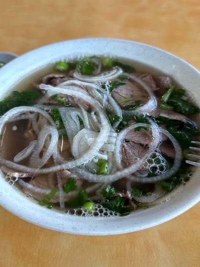A close-up, overhead shot shows a bowl of pho, a Vietnamese noodle soup, resting on a light brown wooden table. The bowl is white with a subtle speckled texture around the rim. Inside the bowl, a clear broth is filled with thin slices of cooked beef, white onion rings, and chopped green onions and cilantro. The soup is steamy, with some small bubbles visible on the surface. A fork is partially visible on the right side of the bowl. The background is out of focus, showing the continuation of the wooden table.