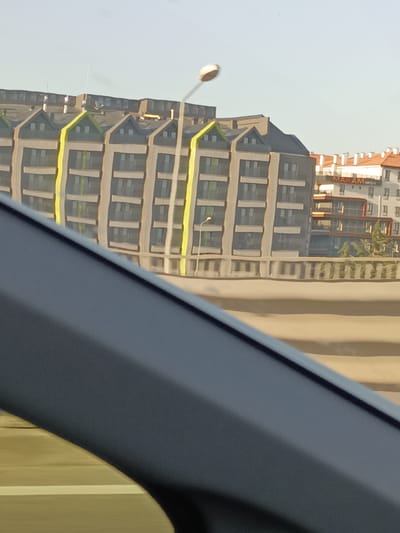 The image shows a distorted reflection of several buildings, possibly due to a watery surface or curved glass. In the foreground, a dark, curved object, possibly part of a car interior, obscures the bottom left of the frame.

The reflected buildings are modern and appear to be apartment or office blocks. The main building reflected on the left has a distinctive grey facade with prominent yellow vertical accents and green triangular roof details. It features multiple floors with regularly spaced windows. To the right of this, a white building with red-tiled roofs is visible. This building has balconies and several windows. Text on this building reads "MAKAM OKCLU".

The scene is set outdoors, under a bright, clear sky, suggesting daytime with fair weather. The distortion of the reflection implies an unusual viewing angle or surface. The overall impression is a somewhat surreal and abstract representation of urban architecture. The specific location context of Yahşihan, Turkey, is provided.