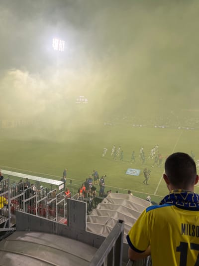 The image depicts a nighttime scene at the Green Stadium in Nof HaGalil, Israel, viewed from an elevated spectator stand. The football field is blanketed by a dense, yellowish-green smoke or haze, significantly reducing visibility. Bright stadium floodlights, including a prominent array on the upper left, pierce through the smoke. A digital scoreboard, partially obscured by the haze, displays "00:00" and scores of "0 0" in red numerals.

Numerous individuals are present on the field. These include groups of athletes in white uniforms and dark green or blue uniforms, dispersed across the turf. Other individuals in darker clothing, consistent with staff or media personnel, are visible closer to the stands. One individual on the field appears to be holding a small child. A blue mat with a logo is positioned on the field.

In the lower right foreground, the back of a male spectator's head and torso are visible. He has short hair and wears a yellow short-sleeved shirt with blue trim on the collar and sleeves. The back of his shirt features the name "MILSO" and the number "17." He is oriented towards the field. Below the spectator's position, tiered rows of empty, folded white seats and metal barriers are visible.