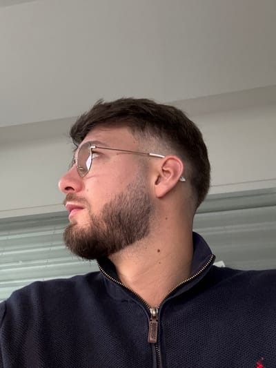A close-up, profile image depicts an adult male from the chest upwards, facing towards the viewer's left. The male has short, dark brown hair with faded sides, a trimmed dark brown beard, and is wearing round, gold-rimmed glasses with tinted lenses and white temple tips. A faint linear scar is visible on his left cheek, and a small white object, possibly an earbud, is inserted into his right ear. He wears a navy blue, textured knit, zippered jacket with a metal zipper and a brown leather pull tab. The subject's head is slightly angled upward, and his gaze is directed out of frame. The background consists of a plain light grey ceiling or wall in the upper portion and a horizontally ridged, light grey or metallic surface, possibly a garage door or storage unit, in the lower portion. There are no identifiable features within the image to confirm a specific location in Lièpvre, France.