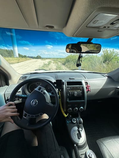 The image is a first-person perspective from inside a car, looking forward. The driver's hands are visible on the steering wheel, which has a Nissan logo. The dashboard features a radio, climate controls, and a gear shift. A rearview mirror reflects the road ahead, which appears to be a dirt track in a rural area with greenery and a blue sky with clouds. In the distance, a large structure, possibly a wind turbine, is visible on the left. The car is likely located on the outskirts of Abrykosivka, Ukraine, given the presence of rural landscape features.