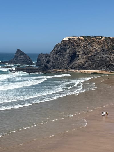 This is a photograph of Odeceixe beach in Odeceixe, Portugal. The image captures a wide view of the beach and ocean under a clear blue sky. In the foreground, gentle waves are washing ashore on a sandy beach. Further out, the ocean is a deep blue with white surf breaking around dark, rocky outcrops and larger sea stacks that rise from the water. Behind the ocean and rocks, a large, rugged cliff face dominates the background, covered in sparse vegetation and showing signs of erosion. On the beach, a few small figures are visible. Two individuals are walking together near the water's edge, one carrying a surfboard. Another individual is walking further up the beach. Small red and white flags are planted in the sand along the waterline. The scene appears to be a sunny day, likely during a time of calm weather at sea.