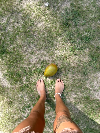 The image shows a person's feet standing on a patch of sandy ground with sparse green grass and a few small plants. A single green coconut is positioned between the person's feet. The person has a tattoo on their right thigh and is wearing a white beaded anklet on their left ankle. The ground appears to be a natural outdoor setting, possibly a beach or park area in Ban Khlong Hia, Thailand, given the presence of sand and vegetation. The lighting suggests it is daytime.