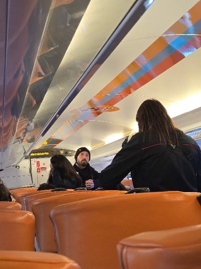 The image captures the interior of a public transport vehicle, identified as being in Cap-d'Ail, France. The perspective is from a lower vantage point within the passenger cabin, looking forward. Numerous orange, upholstered seats are arranged in rows, occupying the foreground and mid-ground.

Three individuals are visible in the mid-ground. On the right side, a person with long, dark hair is seated with their back to the viewer, wearing a black jacket accented with red trim on the shoulders and a dark shoulder strap. To their left, a male subject with a beard and mustache is seated, wearing a black beanie and a dark jacket; he is looking directly towards the camera/viewer, with his hands clasped in front of him. Immediately to the left of the male, another individual with long, dark hair is seated, resting their head on their hand.

The ceiling of the vehicle is white, featuring prominent horizontal striped patterns in orange, light blue, and a darker blue/purple. These stripes are interspersed with reflective, possibly metallic, panels. Integrated overhead lighting fixtures provide illumination. On the upper left side of the cabin, a white sign with black text clearly reads "Zone des départs" (Departure Zone), with a red circular symbol positioned directly below the text. The overall scene depicts passengers in a train or tram environment.