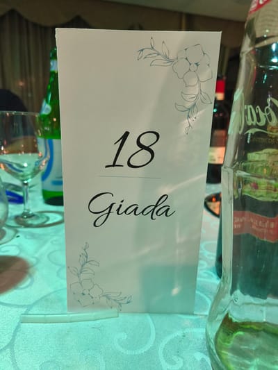 A table number card, designated "18" and bearing the name "Giada" in elegant script, stands upright on a patterned tablecloth. The card features delicate floral designs in the upper right and lower left corners. To the left of the card, a wine glass and a green bottle are visible. To the right, the distorted reflection of a bottle, possibly a Coca-Cola bottle, is apparent. In the background, a curtain hangs, and other bottles and glasses are dimly lit. The lighting suggests an indoor setting, possibly a restaurant or event venue. The image does not provide specific information about the city of Vinchiaturo, Italy.