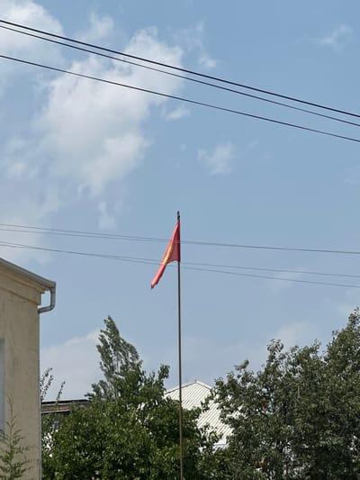 The image depicts a bright, slightly overcast day in the city of Kadamjay, Kyrgyzstan. In the center, a red flag with a yellow emblem is prominently displayed on a tall flagpole. The flag appears to be made of fabric and is slightly rippled by the wind. To the left, a portion of a light-colored building with a grey gutter is visible, suggesting an urban or semi-urban setting. In the background, dense green trees frame the lower part of the scene, with their leaves rustling. Several thin, dark power lines stretch horizontally across the sky, partially obscuring the blue sky and white clouds. The overall impression is one of a typical day in a residential or commercial area of Kadamjay.