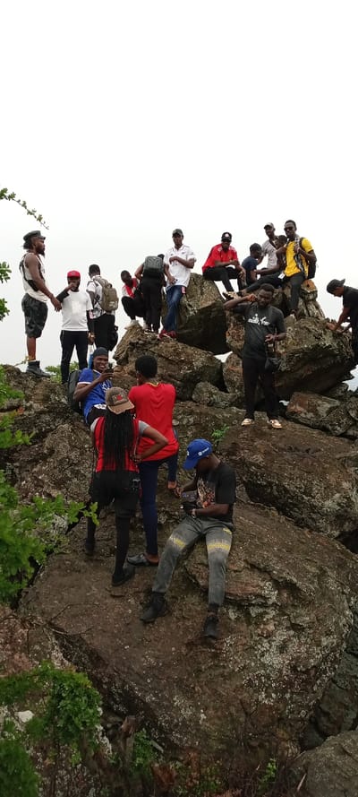 The image captures approximately 15-18 individuals, predominantly young adults of Black ethnicity, gathered on a rocky outcrop in Girei, Nigeria. The scene is outdoors under an overcast sky, featuring a landscape of large, irregular boulders and rocks, some with visible moss or lichen. Sparse green vegetation is present in the lower left foreground.
Individuals are distributed across the uneven terrain at varying elevations. In the left foreground, a man with dreadlocks stands, wearing a white tank top, dark shorts, and dark boots. Below and to his right, a man in a blue t-shirt and backpack is seated on a rock, holding a smartphone and looking towards the camera with a smile. Behind him, a woman with long braids, wearing a red top, dark shorts, and leggings, stands facing away from the camera, hands on her hips. In the lower right foreground, a man in a black t-shirt, blue baseball cap, and grey patterned pants is seated on a rock, looking down at a small object in his hands.
Towards the center and background, other members of the group are positioned on the rocks. A man in a black t-shirt stands with a camera hanging from a strap around his neck. Higher on the rocks, individuals are seen standing and sitting; their attire includes a white Arsenal FC jersey, a red Manchester United FC jersey, various t-shirts (yellow, black, white), and baseball caps. Several individuals are wearing backpacks. The collective actions suggest a group outing or hike, with individuals resting, observing, and engaging with personal devices.