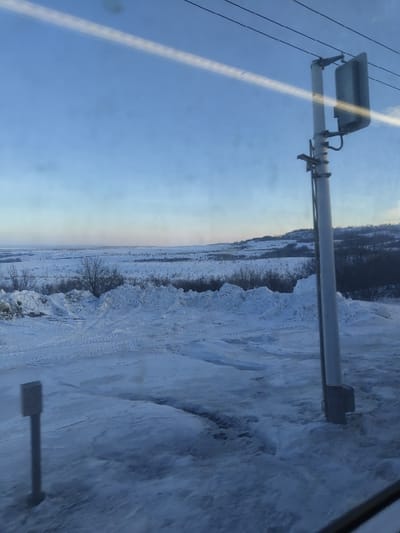 The image presents a snowy, open landscape viewed from inside a vehicle, likely through a window given the prominent reflections. The overall impression is one of a cold, winter day.

In the foreground, on the left side of the frame (when oriented correctly), a tall, light-colored utility pole or streetlight stands. Attached to its upper portion is a light fixture or a piece of equipment, and wires extend horizontally from it towards the left edge of the image. A strong, diagonal streak of bright light, possibly a reflection from the window, cuts across the sky from the upper left to the lower right. Other fainter smudges and reflections are also visible on the window surface. Another, shorter, darker post or marker is partially visible in the bottom right corner of the frame, emerging from the snow.

The ground is heavily covered in snow, showing varying textures and drifts, suggesting uneven terrain. Faint tracks, possibly from a vehicle or animal, can be discerned in the snow, winding across the landscape. In the midground, beyond the immediate snowy expanse, there are hints of low-lying vegetation or brush, also covered in snow.

The sky occupies the upper portion of the image. It is a clear blue, transitioning to a lighter, almost hazy, tone near the horizon, suggesting either early morning or late afternoon light. In the far distance, a faint, dark line indicates a horizon with possibly distant hills or a treeline.

The photograph captures a static scene, but the nature of the reflections and the distant landscape hint that it was taken from a moving vehicle, possibly a train or car, traveling alongside the snowy environment. There is no visible text in the image.