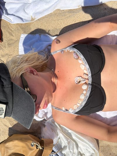 A woman is lying on a sandy beach on top of two white towels. She is wearing a black bikini top with "Calvin Klein" printed on the straps and has seashells arranged on her chest and upper abdomen. A silver necklace is visible around her neck. She is wearing sunglasses with a gold logo and a gray baseball cap with a patch. A tan leather drawstring bag is partially visible to her right. The background consists of light brown sand, indicating a beach setting. The location of Toseong-myeon, South Korea, is not evident in the image.