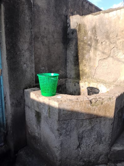 This image captures an outdoor corner scene in Njoro ward, Kenya, featuring weathered concrete structures under direct sunlight. A bright green, perforated plastic wastebasket stands centered on a raised, rectangular concrete ledge. To the immediate right of the basket, the concrete structure forms a cuboid recess, within which a distinct shadow of the basket is cast. The two concrete walls forming the corner behind the ledge exhibit extensive discoloration, dark staining, and structural cracking. A sharp, diagonal shadow bisects the right concrete wall and the upper surface of the concrete structure. A narrow, faded blue door frame is visible along the far left edge of the frame. A patch of clear blue sky is discernible above the right wall, which also displays several small plant sprouts along its top edge. No individuals or active interactions are present in the scene.