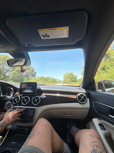 This is a first-person point of view, indoor shot of the interior of a car, likely a Mercedes-Benz, in Ruska Bela, Bulgaria. The view is out the windshield, showing a clear, blue sky and a lush, green landscape with trees and rolling hills. The dashboard is visible, featuring a central infotainment screen displaying "Bluetooth Audio Device Paused," and circular air vents. A person's hand is seen reaching towards the car's audio controls, holding a smartphone. The legs of two people are visible in the foreground, one with tattoos on their thigh and calf. A pink fuzzy air freshener hangs from the rearview mirror. The overall impression is a snapshot of a car journey on a sunny day.