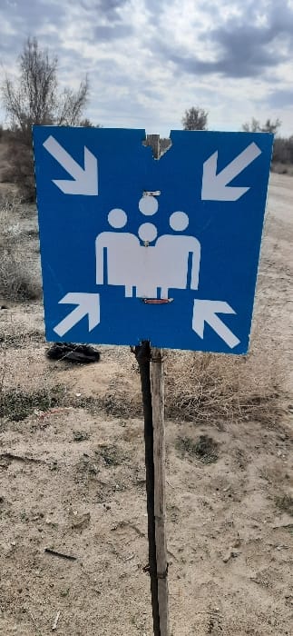 An upright, blue rectangular sign, approximately 2 feet by 2 feet, displaying white graphics signifying an assembly point, is centrally positioned outdoors in an undeveloped area within or near Qorovulbozor, Uzbekistan. The sign features four white arrows pointing inwards towards a central image of three stylized white human figures, consisting of two larger figures and one smaller figure positioned centrally and slightly above them. A small, closed red pocket knife is affixed horizontally to the sign below the figures. The sign exhibits a V-shaped tear at its top center and multiple small circular marks (approximately 0.5 cm in diameter) near the upper part of the graphic. It is mounted to a rough, vertical wooden pole, which is partially wrapped with dark material. The pole is embedded in sandy, uneven ground covered with sparse, dry, light-brown vegetation and scattered debris, including a dark irregular object to the left of the pole. In the midground and background, several leafless, bushy trees with thin branches are visible under an overcast sky with grey clouds. No people or active interactions are observed in the scene.