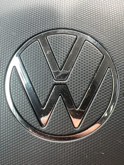 The image shows a close-up, detailed view of the Volkswagen logo. The logo is circular with a metallic finish and is embossed on a textured black surface that resembles a honeycomb pattern. The logo itself is a stylized "VW" within the circle, with the "V" positioned above the "W". Reflections are visible on the metallic surface of the logo, indicating it is likely a car emblem or part of a car's interior. The background texture is consistent and appears to be a durable material. There are no people or other objects visible in the frame. The lighting suggests an indoor environment, possibly artificial light. The image's focus is sharp on the logo and the immediate surrounding textured surface. The location in Siegen, Germany is not discernible from the visual information provided in the image.