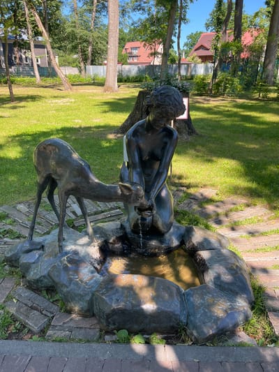 This media file features a bronze fountain sculpture of a male figure and a deer. The sculpture is situated in an outdoor setting with lush green grass and mature trees. Sunlight casts dappled shadows on the ground, suggesting it is daytime, likely late morning or early afternoon, with clear skies and fair weather. The background shows residential buildings with red-tiled roofs, hinting at a park or residential area. The sculpture depicts the male figure reclining, interacting with the deer, which appears to be drinking from the fountain. Water is actively flowing from the sculpture, creating a refreshing atmosphere. The sculpture itself is detailed, with the bronze showing signs of patina and age. The overall scene evokes a sense of tranquility and natural harmony.