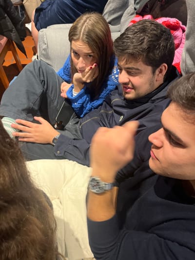 Three people are sprawled on a couch covered with blankets. A woman in a blue sweater looks up with a hand near her face. Beside her, a man with dark hair rests his head. Another man, wearing a navy sweater, is to the right. He wears a watch, and his arm is extended towards the camera, slightly out of focus. A cell phone and small bottle sit on the couch near the center. Part of a wooden chair and brown hair are visible in the lower part of the frame.