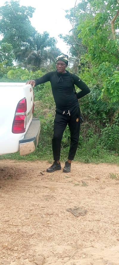 A man is standing near a white pickup truck on a dirt road in Otunade, Nigeria. He is wearing a black long-sleeved shirt, black sweatpants, black shoes, and a camouflage-patterned head covering. He is leaning against the truck with his left hand resting on the truck bed, and his right hand on his hip. The surrounding environment includes trees and vegetation.