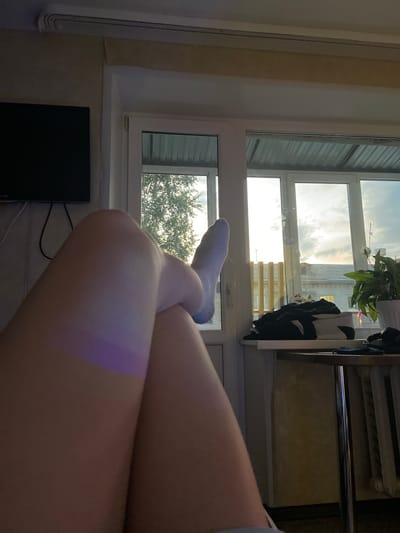 The image shows a person's legs in the foreground, crossed, with one foot wearing a light blue sock. A purple light is cast on the left leg. In the background, a window with multiple panes reveals an outdoor scene with trees and buildings under a cloudy sky. A television is mounted on the wall to the left. A shelf on the right holds several folded clothes and a potted plant. The overall setting appears to be an indoor living space with a view of an urban environment. The city of Beloretsk, Russia, is mentioned in the prompt but there are no visual elements in the image that confirm this specific location.