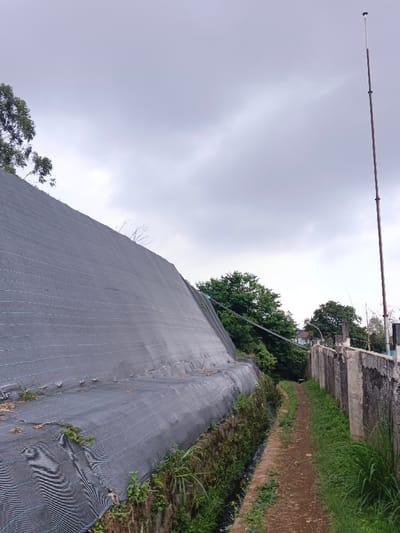 The scene depicts an outdoor, unpopulated pathway in Koipa, Papua New Guinea, under an overcast sky. On the left, a steep earthen embankment is covered by black woven mesh, consistent with geofabric or shade cloth, secured horizontally with lines and extending upwards and into the background. Sparse green vegetation grows at the base of this slope, adjacent to a narrow, dark drainage channel. A dirt path, approximately 1.5 meters wide, occupies the middle ground and recedes into the distance. To the path's right, a weathered concrete block retaining wall runs parallel, displaying exposed aggregate and dark staining. Beyond this wall, dense green foliage and trees are visible. A tall, slender, segmented pole, consistent with a utility or antenna mast, stands vertically on the far right, extending towards the sky. Multiple tensioned cables traverse the upper mid-ground, extending from the vicinity of the covered slope towards the right side of the frame.