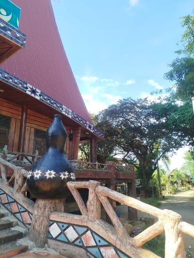 The image depicts a building in An Phu Ward, Vietnam. The building has a red corrugated metal roof and a wooden facade with a decorative trim featuring geometric patterns. The building is elevated with a staircase leading to the entrance. The staircase has a textured, faux-wood railing decorated with geometric tile patterns. A large, gourd-shaped, black-and-white patterned object sits atop one of the railing posts. In the background, a large tree with dense foliage is visible, along with a glimpse of a street or path and other buildings. The sky is blue with scattered clouds. The scene is well-lit, suggesting daytime.