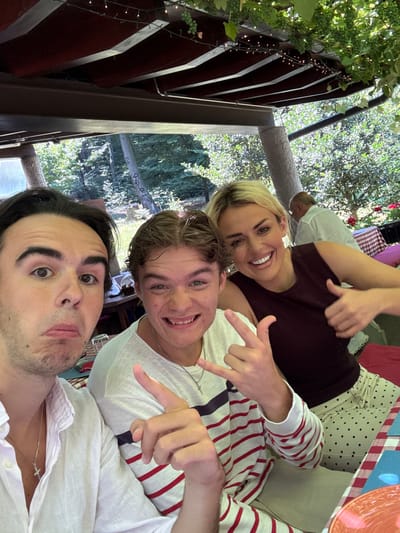 In the city of Agno, Switzerland, three individuals are captured in a selfie outdoors under a wooden pergola adorned with string lights and grapevines. The man on the left, with dark hair and a surprised expression, wears a white button-down shirt and a silver cross necklace. Beside him, a young man with light brown hair grins widely, making a hand gesture, and sports a white t-shirt with red stripes. To the right, a blonde woman with a bright smile gives a thumbs-up, wearing a dark sleeveless top and polka-dot pants. In the background, a man in a white shirt is visible, and red and white checkered tablecloths and cushions suggest a dining setting. The environment appears to be a restaurant or a garden patio, with lush green trees and foliage surrounding the area.