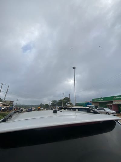 This image captures a street scene in Masii ward, Kenya, viewed from the interior of a white vehicle, likely an SUV, with its roof, roof rack, and antenna visible in the foreground. The sky is heavily overcast with dense grey clouds; two small, dark birds are in flight high above.
The midground depicts a busy road with numerous vehicles and pedestrians. On the left side of the road, multiple utility poles with a dense network of overhead wires line what appears to be a market area with several small, indistinct structures or stalls. Several individuals are visible, including one person wearing a yellow hard hat standing near the center of the road, partially obscured by other vehicles. On the right side of the road, a prominent green building with white lettering displays "Safaricom m-pesa" signage, indicating a commercial establishment. A silver SUV is parked or slowly moving in front of this building. A tall, multi-lamp floodlight pole stands prominently in the center-right, extending high into the cloudy sky. Other vehicles, including a white truck and various cars and vans, are present on the road, suggesting active traffic or congestion. The overall environment appears to be an urban or semi-urban commercial thoroughfare.