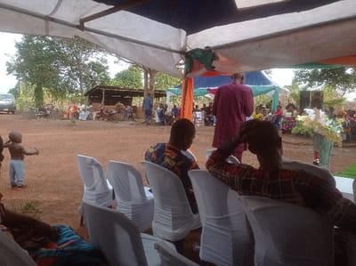 The scene depicts an outdoor gathering in Ugbongha, Nigeria, under a series of temporary tarpaulin and fabric shelters. The ground is reddish-brown bare earth.
In the foreground, a white tarp roof is visible, supported by a wooden post on the left. Draped beneath the tarp are sections of green, orange, and blue fabric. Two adult males are seated with their backs to the camera on white chairs covered in white fabric. The man on the left wears a patterned shirt, and the man on the right wears a red and black plaid shirt, with his right hand resting on his head. To their left, a young child, shirtless, wearing light blue pants and red shoes, stands facing left, holding a thin stick with both hands. A portion of a person's legs covered by a colorful patterned fabric is visible at the bottom left.
In the midground, a man in a maroon long-sleeved garment stands with his back to the camera, addressing an audience. The audience, composed of multiple individuals mostly seated on chairs under a light-colored canopy (possibly green or white), faces the speaker. Many women in the audience wear colorful attire. To the right of the audience, a large black speaker rests on the ground next to a decorative green woven vase containing white artificial flowers and green foliage. Further back, near a simple wooden shed with a dark roof, a person in blue attire walks left.
The background features sparse trees and bushes. A white vehicle is partially visible behind vegetation on the far left. Near the wooden shed, two motorcycles are parked alongside stacks of items in sacks and bundles. The sky is overcast.