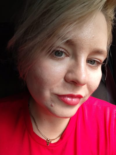 A close-up, eye-level, medium shot shows the face of a young blonde woman wearing a bright red shirt and a gold chain with a small cross pendant. Her short, tousled blonde hair is partially obscuring her forehead and the top of her right eye. She has fair skin with visible pores and a slight sheen, suggesting a natural or lightly made-up look. A small reddish blemish is visible on her chin, just below her lower lip.

Her eyes are open and looking slightly to her left, with light blue irises and dark eyelashes. She is wearing subtle eye makeup. Her lips are full and painted with a vibrant pink lipstick that has a glossy finish, giving them a plump appearance. A subtle smile plays on her lips, conveying a sense of confidence and playfulness.

The background is dark and out of focus, with a hint of a black seat visible on the right side of the frame, suggesting she might be in a car. The lighting appears to be natural, possibly from a window, illuminating her face from the left and creating soft shadows. The overall mood is casual and perhaps a bit candid, as if she is taking a selfie. There is no visible text in the image. The location context of Vorob'evo, Russia, does not provide any specific visual cues in this close-up shot.
