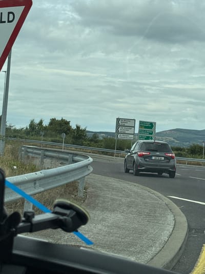 The image shows a grey Toyota Yaris driving on a road. The car has a license plate that reads "201-D-611". In the background, there are several road signs. One sign indicates directions to "Borrris", "KILKENNY", and "MOUNTIELLOT". Another sign points towards "ENNISCORTHY", "CLONROCHE", and "DUBLIN". The sky is overcast and there are hills in the distance. The location appears to be in or near New Ross Municipal District, Ireland, as indicated by the road signs.