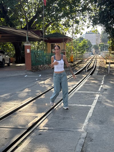 A female stands on a narrow-gauge railroad track in Lao Cai Ward, Vietnam. She is wearing a white tank top and light-wash jeans. The track is embedded in a concrete surface. Vegetation is visible on the left, including a large tree and green fencing. Buildings can be seen in the distance, along the line of the track. The scene is outdoors in daylight.