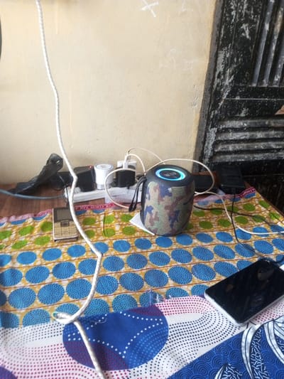 A table covered with a colorful patterned cloth displays several electronics. A camouflage-patterned speaker with a glowing blue top sits near a white multi-outlet power strip crammed with various chargers. A black smartphone lies on the right side of the table, and a small brown flip phone is visible on the left. White cables snake across the surface, connecting devices to the power strip. A partially visible weathered door stands behind the table.