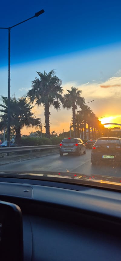 The image captures a sunset scene in Alimos, Greece, viewed from inside a vehicle. The sky displays a gradient of deep blue at the top, transitioning to lighter blue and then to a warm orange and yellow hue near the horizon where the sun is setting. Palm trees, characteristic of coastal Mediterranean locations like Alimos, line the right side of the road, silhouetted against the vibrant sky.
The foreground shows the interior of a car, with the dashboard visible. The road ahead is lined with a metal guardrail. Several cars are driving on the road. The closest vehicle in the right lane has a license plate that reads "I EX 1846". To its left, another car with a license plate "BKA 6397" is also visible. Further down the road, more vehicles and palm trees are present, with the setting sun casting a warm glow. A street lamp is prominently visible on the left side of the frame, extending upwards into the blue sky. The overall impression is one of traffic moving along a coastal road during a beautiful sunset.