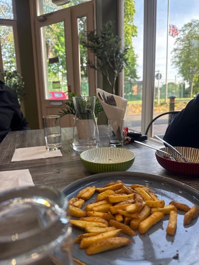 The image displays an indoor dining scene, likely within a cafe or restaurant, taken from a low, slightly angled perspective across a table. The primary subject in the mid-ground is a plate of golden, seasoned fries (chips) resting on a grey ceramic plate with a textured rim.

On the wooden-effect table, other items include a red and a green striped bowl, a glass pitcher, a smaller drinking glass, and a napkin dispenser holding cutlery. A set of keys with a small decorative figure is also visible on the table. A person in a dark top is partially visible, seated at the table, with their arm and torso in the upper left and their lower body in the lower right.

The background is dominated by large windows, allowing ample natural light into the space, indicating it is daytime. Through the windows, green trees and a bright sky are visible. Notably, a Union Jack flag is clearly seen flying in the distance, reflected in the upper window panes, confirming the location as the United Kingdom (specifically Rushmoor, as hinted). A red vehicle, possibly a truck or bus, can be faintly observed through the windows in the lower distance. Green plants, possibly decorative foliage, are placed near the windows, enhancing the bright and airy atmosphere. The scene conveys a casual meal setting. No legible text is visible in the image.