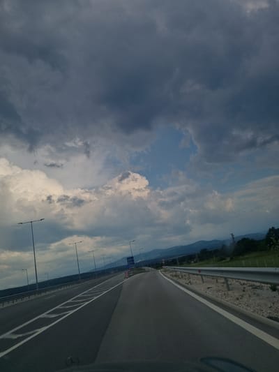 The image depicts a highway under a dramatic sky filled with dark, heavy clouds and patches of bright sunlight breaking through. The highway appears to be an elevated section or a bridge, with guardrails on the right side and a series of streetlights lining the left. The road has multiple lanes with white markings. In the distance, a range of hills or mountains is visible, covered in trees. The overall impression is one of an approaching storm or a moody, overcast day.

The specific location is identified as the highway near Dimovo, Bulgaria, likely a stretch of the European route E79, which passes through the region. There are no people visible in the image. The essential objects include the highway infrastructure: asphalt road, white lane markings, metal guardrails, and tall streetlights. The environment is characterized by the sky and the distant mountainous landscape. No actions or interactions are occurring.