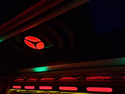 The image presents an interior view with futuristic or stylized lighting, seemingly within a bus or similar transportation vehicle. The lighting scheme is dominated by red and green hues. Red neon lights are visible as horizontal strips, illuminating the lower portion of the scene. Above them, a band of green light is present. The ceiling has a dark background with a concentric circle pattern around a central red three-pronged shape. There is no evidence of people, and the focus remains on the lighting and interior design elements. There is no location information to verify Anipemza, Armenia.