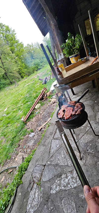 The image shows a barbeque being cooked outdoors. The barbeque grill, round and black, is filled with meat. Smoke rises from the grill. Tongs are held by a hand, positioned over the grill. The ground is a mix of concrete and grass. In the background, there's greenery, a wooden structure, and various objects on a table. The location is Sveti Konstantin, Bulgaria.