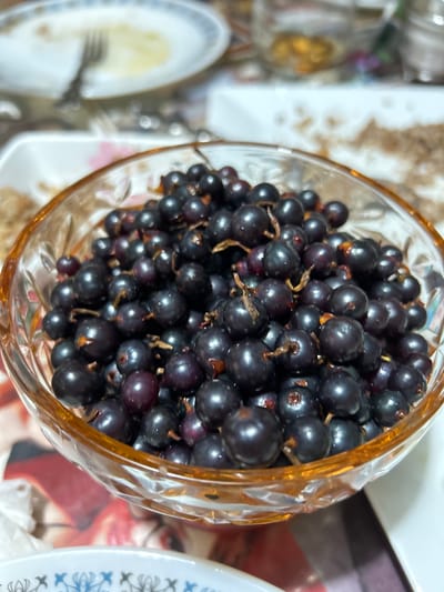 A close-up, eye-level shot showcases a clear, faceted glass bowl filled with ripe black currants. The currants are dark, almost black, with some exhibiting a deep purple hue and small, lighter brown stems. They are densely packed in the bowl, creating a textured surface. The bowl itself has a warm, amber tint around the rim and appears to be made of thick glass, possibly crystal, given the way it catches the light. The background is blurred, suggesting it's a domestic setting. Partially visible in the upper left is a white plate with a fork resting on it, hinting at a meal. Other out-of-focus elements in the background suggest a table setting with more dishes and possibly other food items. The lighting is soft and natural, illuminating the currants and creating subtle highlights on their surfaces. There is no indication of the specific location in Copceac, Moldova, other than the general context of a home environment.