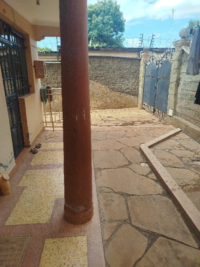 The image provides a forensic view of an outdoor courtyard and building entrance located in Kihara ward, Kenya. A large, reddish-brown cylindrical column dominates the center-left foreground. To its left, a cream-colored building facade features a dark metal security door and a window secured with dark metal bars. The building's porch floor is covered with a pattern of yellow speckled rectangular tiles bordered by reddish speckled tiles. On this porch, a light-colored, cylindrical pot rests near the column, and two dark, irregular objects are positioned closer to the door. A metal railing is visible further back on the porch, with a green cloth draped over it, and a light-colored box is mounted on the building wall.

To the right of the column, the courtyard floor consists of irregularly shaped, light brown flagstones with dark grout, showing evidence of moisture. An 'L'-shaped border of reddish speckled material is embedded in the flagstone paving in the lower right. The courtyard is enclosed by a dark, textured concrete wall exhibiting crack patterns, topped with razor wire and barbed wire. A dark blue/black ornate double-gate, featuring decorative elements, is set within stone pillars topped with spherical finials on the right side of the frame. Beyond the wall and gate, green trees and a partly cloudy blue sky are visible.