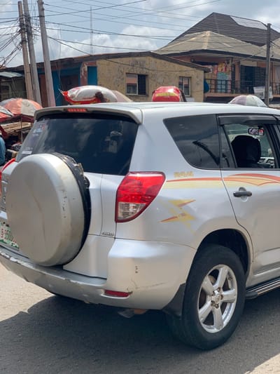 The image shows the rear of a silver Toyota RAV4 parked on a paved road in Ondo, Nigeria (latitude: 7.251, longitude: 5.196). The car is facing the right side of the image, with the rear tire on the left side of the image, and the rear of the vehicle extending to the right. The license plate is partly visible. There is a spare tire cover on the rear hatch, and the words "RAV4" and "4WD" are visible on the rear hatch. There are two red bags secured to the roof of the vehicle. The car is parked near other buildings, a busy street and power lines. The sky is overcast and partially obscured by power lines. There is a yellow and red stripe along the side of the car. There is a reflection in the rear window. There are no people visible in the image.