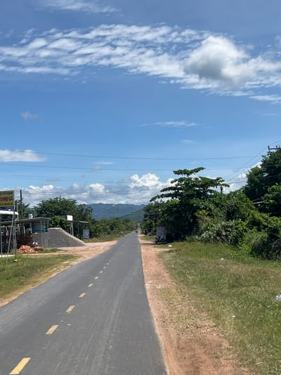 The image depicts a rural road in Ham Thuan Bac, Vietnam, on a partly cloudy day. The road is paved with asphalt and has yellow dashed lines marking the lanes. On the left side of the road, there is a small construction site with stacks of bricks, piles of gravel, and some makeshift shelters. A sign with Vietnamese writing is also visible. On the right side of the road, the terrain is covered with grass and scattered debris. Lush green trees and vegetation line both sides of the road, with rolling hills and mountains visible in the distance. Power lines are strung across the sky. A white motorcycle is parked on the side of the road near the construction area. The sky is a vibrant blue with scattered white cumulus clouds.