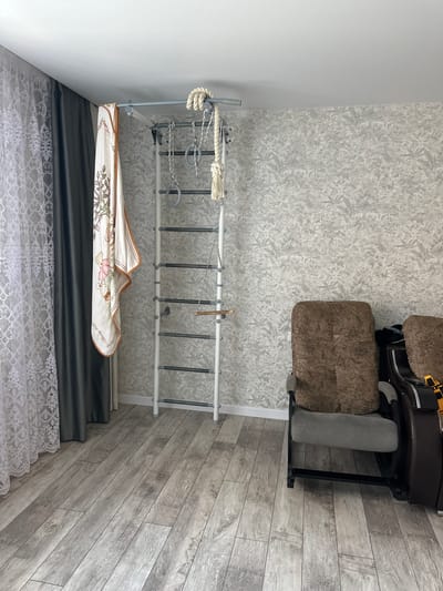 The image depicts an indoor scene, likely a living room or playroom, with a focus on a wall-mounted gymnastics apparatus. The apparatus is white with dark gray rungs and features various attachments, including rings, a rope with a thick knot, and a small wooden plank. A light-colored blanket with a cartoon bear print is draped over one side of the apparatus.
To the left of the gymnastics equipment, there are two sets of curtains: a sheer white lace curtain layered over a dark gray opaque curtain. The wall behind the apparatus is covered in a textured, light gray wallpaper with a subtle botanical pattern.
In the foreground and to the right, a dark brown recliner-style chair is partially visible. The chair has patterned cushions and appears to be in a reclined position. The flooring is a light gray laminate with a wood plank design. The overall impression is a well-lit, domestic space. The specific location in Mariinsky, Russia cannot be determined from the visual information provided in the image.