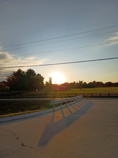 The image depicts a rural landscape at sunset in Paint Lick, United States. The sun is low on the horizon, casting a warm glow over a field with a dark wooden fence. Several trees, including a cluster of dense evergreens, are silhouetted against the bright sky. In the distance, a red barn and a small blue structure are visible. The foreground features a paved road with a metal guardrail, its shadow stretching across the asphalt. The sky is a gradient of pale blue to warm orange, with a few wispy clouds. Power lines are strung across the upper portion of the frame.